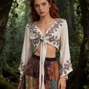 Può includere: Top corto color crema con maniche lunghe e ricami geometrici. Il top è annodato in vita. La modella indossa una gonna patchwork con un motivo colorato e una fascia per capelli fantasia. Lo sfondo è una lussureggiante foresta verde.