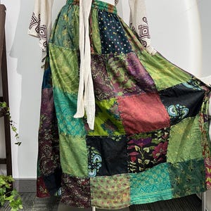 Puede incluir: Una falda patchwork en tonos verdes, morados, rojos y negros. La falda está hecha de varios cuadrados de tela con estampados florales y geométricos. La cintura es verde. La falda se exhibe en un maniquí.