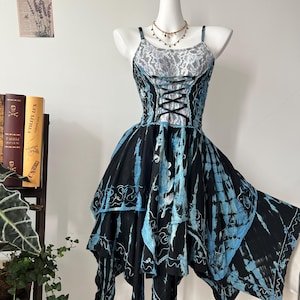 Könnte beinhalten: Ein schwarz-blaues Batik-Kleid mit einem Korsett-Oberteil und Spaghettiträgern. Das Kleid hat einen mehrschichtigen, asymmetrischen Rock und wird auf einer Schaufensterpuppe präsentiert. Eine zarte Halskette ist sichtbar.