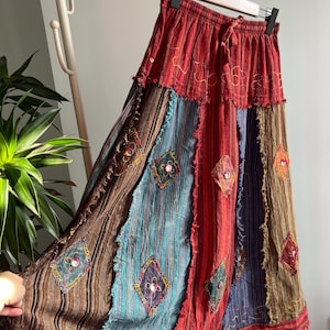 Op de afbeelding: Een lange patchwork rok met een rode, elastische tailleband en trekkoord. De rok heeft verticale panelen van bruine, blauwe en rode stoffen, versierd met geborduurde ruitvormen en kleurrijke stiksels. De rok hangt aan een houten kleerhanger.
