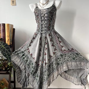 May include: A gray sleeveless dress featuring a lace-up corset-style bodice, a scoop neckline, and a handkerchief hemline. The fabric displays a dark green and red floral pattern with intricate embroidery and a ruffled bottom edge.