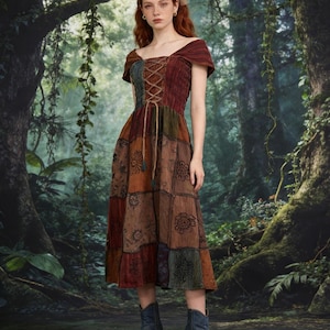 Op de afbeelding: Een patchwork midi-jurk in bruin-, groen- en bourgondische tinten. De jurk heeft een vetersluiting, korte mouwen en een bloemenpatroon op de rok. Het model draagt ​​blauwe enkellaarsjes. De achtergrond is een weelderig bos.