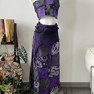 May include: A two-piece set consisting of a strapless tube top and a long wrap skirt. Both pieces feature a patchwork design in shades of purple and black, decorated with printed celestial motifs including suns, moons, and stars with frayed edges.