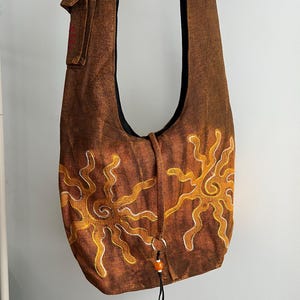 May include: A brown hobo bag featuring two embroidered sun designs in yellow and white thread. The bag has a wide shoulder strap, a small side pocket, and a central drawstring closure accented with a silver ring and a single orange bead.