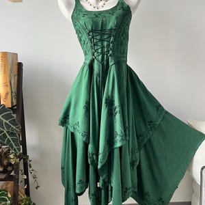 Puede incluir: Un vestido verde con un corpiño estilo corsé y una falda asimétrica. El vestido presenta intrincados bordados florales y un frente con cordones. El vestido se exhibe en un maniquí, mostrando su diseño y estilo.