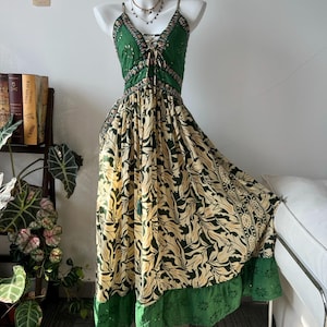 Pode incluir: Um vestido longo sem mangas com um corpete verde-escuro com atacadores e alças finas. A saia apresenta um padrão de folhas em creme e verde-escuro, rematado com uma bainha verde larga com bordados intrincados e recortes.