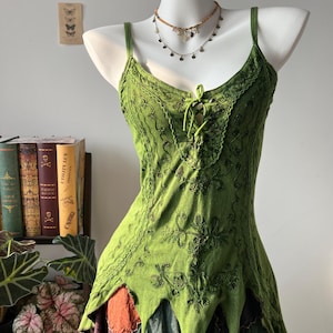 Può includere: Un top verde senza maniche con intricati ricami verde scuro, chiusura frontale con lacci e spalline sottili, e un orlo irregolare a punta. Il capo è sovrapposto a una gonna patchwork con pannelli di tessuto arancione, viola e verde scuro.