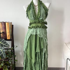 May include: A sleeveless halter-neck dress in various shades of green, featuring a deep V-neckline, frayed fabric edges, and layered textures. The bodice includes ruffled detailing, while the skirt consists of multiple panels of fabric.