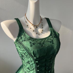 May include: A green dress with a corset-style bodice and embroidered detailing. The dress is displayed on a mannequin and is paired with two gold necklaces with star and moon charms.