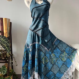 Puede incluir: Vestido halter turquesa con falda patchwork. El corpiño tiene un aspecto texturizado con costuras oscuras. La falda presenta un patrón geométrico en tonos azules y verdes, ribeteado con encaje blanco. Un collar adorna el maniquí.
