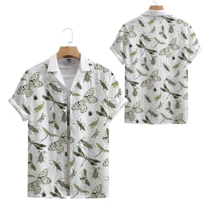 Puede incluir: Camisa blanca de manga corta con cuello de campamento, estampada con insectos y mariposas verdes y negras. La camisa se muestra en una percha de madera. La camisa es de tela ligera.