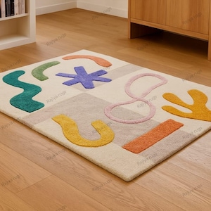 May include: A rectangular area rug with a cream-colored background and colorful abstract shapes. The shapes include a teal squiggle, a purple star, and a yellow U-shape. The rug is on a wooden floor.