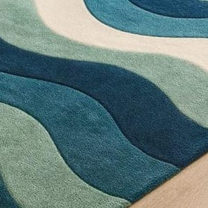 May include: A rectangular rug with a wave pattern in shades of blue, teal, and white. The rug has a soft, textured appearance, with the wave design creating a sense of movement. The rug is placed on a light-colored wooden floor.