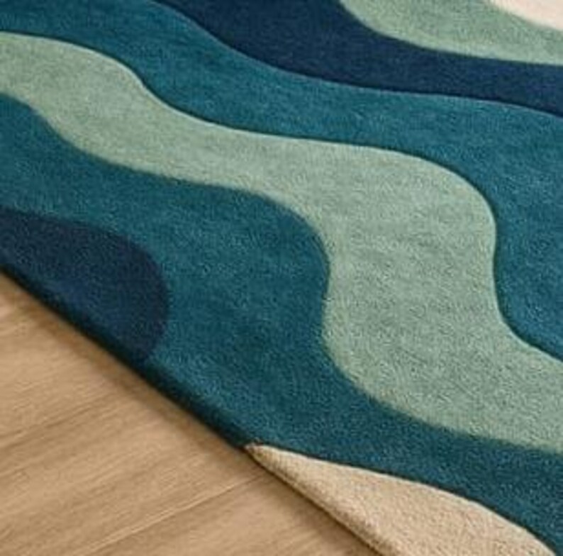May include: A close-up of a rug with a wavy pattern in shades of blue and green. The rug's design features flowing, organic shapes, creating a textured appearance. The rug is placed on a light-colored wooden floor.