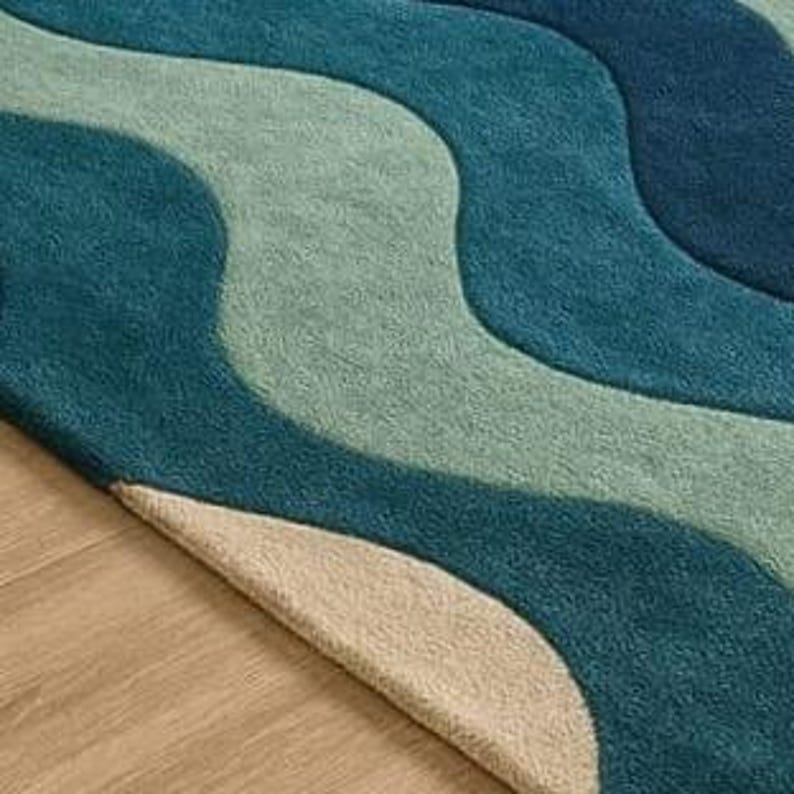May include: A close-up of a rug with a wavy pattern in shades of blue and green. The rug's design resembles ocean waves, with a beige border. The rug is placed on a wooden floor.