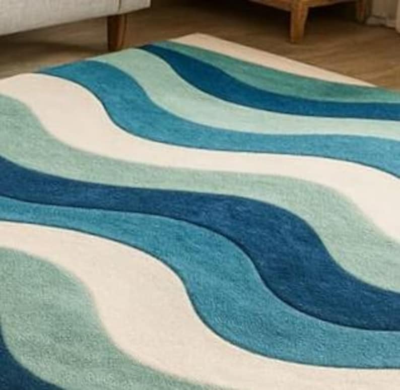 May include: A rectangular rug with a wavy pattern in shades of blue, teal, and off-white. The rug's design features broad, flowing stripes that create a sense of movement and texture. The rug is made of a soft, plush material.