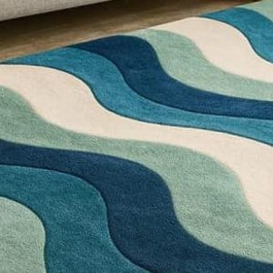 May include: A close-up of a rug with a wavy pattern in shades of blue, teal, and white. The rug has a soft, textured appearance, with the waves flowing across the surface. The rug is a home decor item.