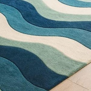 May include: A close-up of a rug with a wave pattern in shades of blue, teal, and off-white. The rug has a soft, textured appearance and is placed on a light-colored wooden floor.