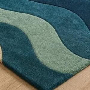 May include: A close-up of a rug with a wave pattern in shades of teal, blue, and green. The rug has a soft, textured appearance and is placed on a wooden floor. The design features flowing, curved lines.