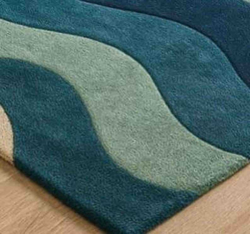 May include: A close-up of a rug with a wave pattern in shades of teal, blue, and green. The rug has a soft, textured appearance and is placed on a wooden floor. The design features flowing, curved lines.