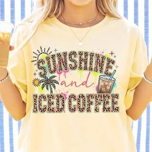 以下が含まれることがあります： ヒョウ柄の「SUNSHINE and ICED COFFEE」という文字がプリントされた淡い黄色のTシャツ。デザインにはヤシの木、太陽、アイスコーヒーのグラスが描かれており、カラフルなペイントの飛び散りと小さな星がアクセントになっています。