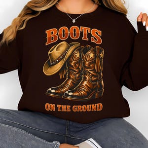 May include: Brown sweatshirt with the text "BOOTS ON THE GROUND" in orange. The graphic features a cowboy hat and a pair of brown cowboy boots with star details. The boots have fringe and are detailed with shading.
