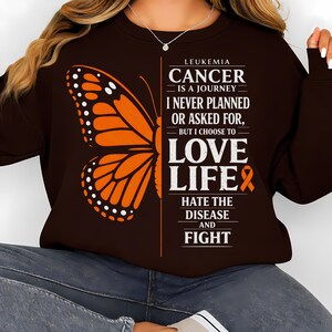 Könnte beinhalten: Ein braunes Sweatshirt mit einem orangefarbenen Schmetterlingsdesign auf der linken Seite. Auf der rechten Seite des Sweatshirts steht der Text "LEUKÄMIE KREBS IST EINE REISE, DIE ICH NIE GEPLANT ODER GEFRAGT HABE, ABER ICH WÄHLE ES, DAS LEBEN ZU LIEBEN, DIE KRANKHEIT ZU HASSEN UND ZU KÄMPFEN."