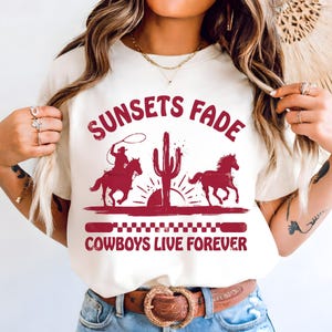 May include: Cream-colored t-shirt with a maroon graphic featuring a cowboy on horseback roping, a cactus, and a running horse. The text reads "SUNSETS FADE COWBOYS LIVE FOREVER".