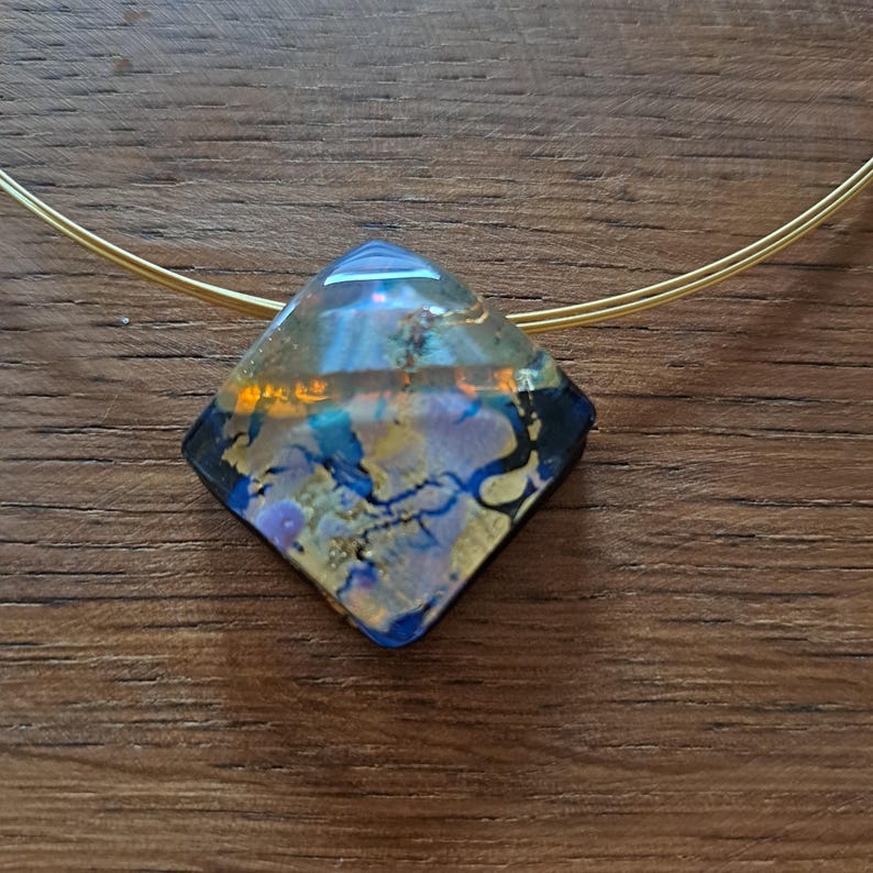 May include: A gold chain necklace with a diamond-shaped pendant. The pendant is made of clear glass with blue, gold, and purple swirls.