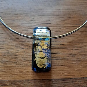 May include: A rectangular pendant necklace with a silver-colored wire. The pendant is a mix of black, gold, and blue glass. The gold has a crackled appearance. The necklace is on a wooden surface.
