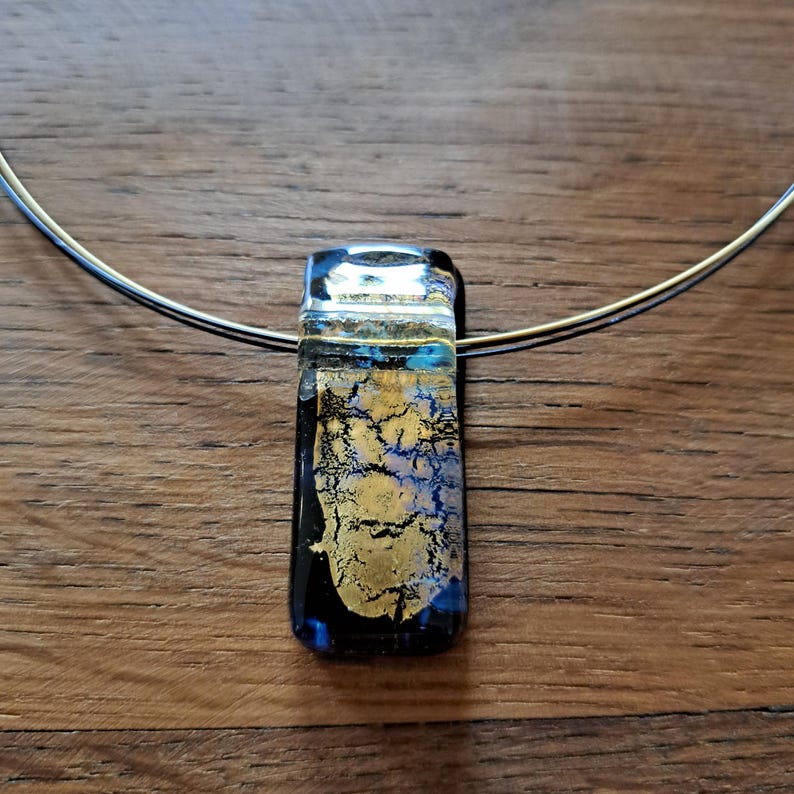May include: A rectangular pendant necklace with a silver-colored wire. The pendant is a mix of black, gold, and blue glass. The gold has a crackled appearance. The necklace is on a wooden surface.