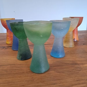 May include: Six small, colorful glass cups with a flared top and a narrow base. The cups are arranged in a semi-circle on a wooden surface.