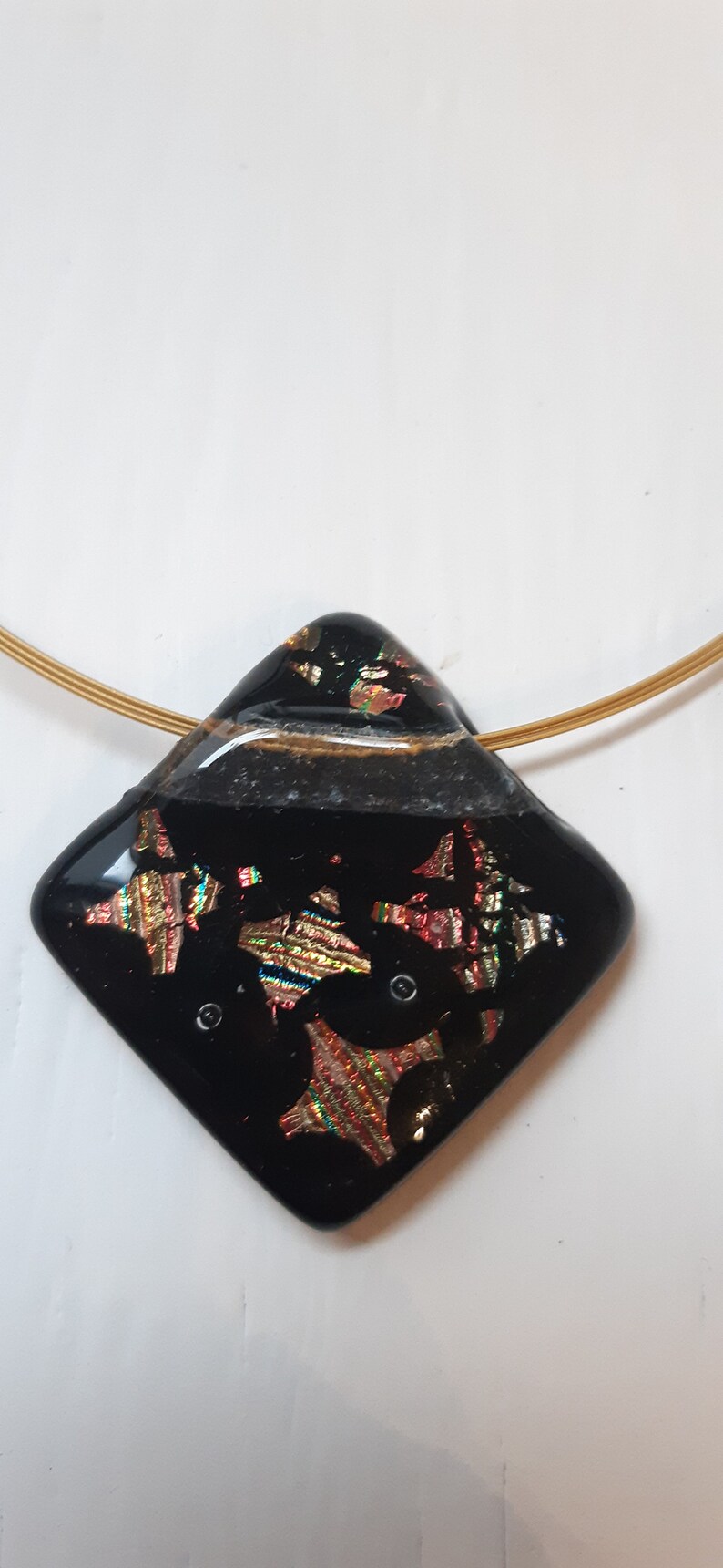May include: A black glass pendant with gold and iridescent accents hangs from a gold chain. The pendant is shaped like a diamond and has a textured surface.