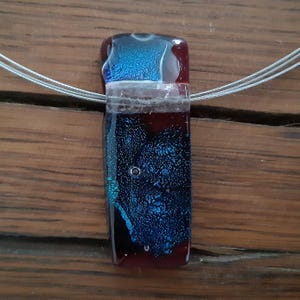 May include: A dark blue and red glass pendant with a silver wire necklace. The pendant has a textured surface and a clear layer in the middle.