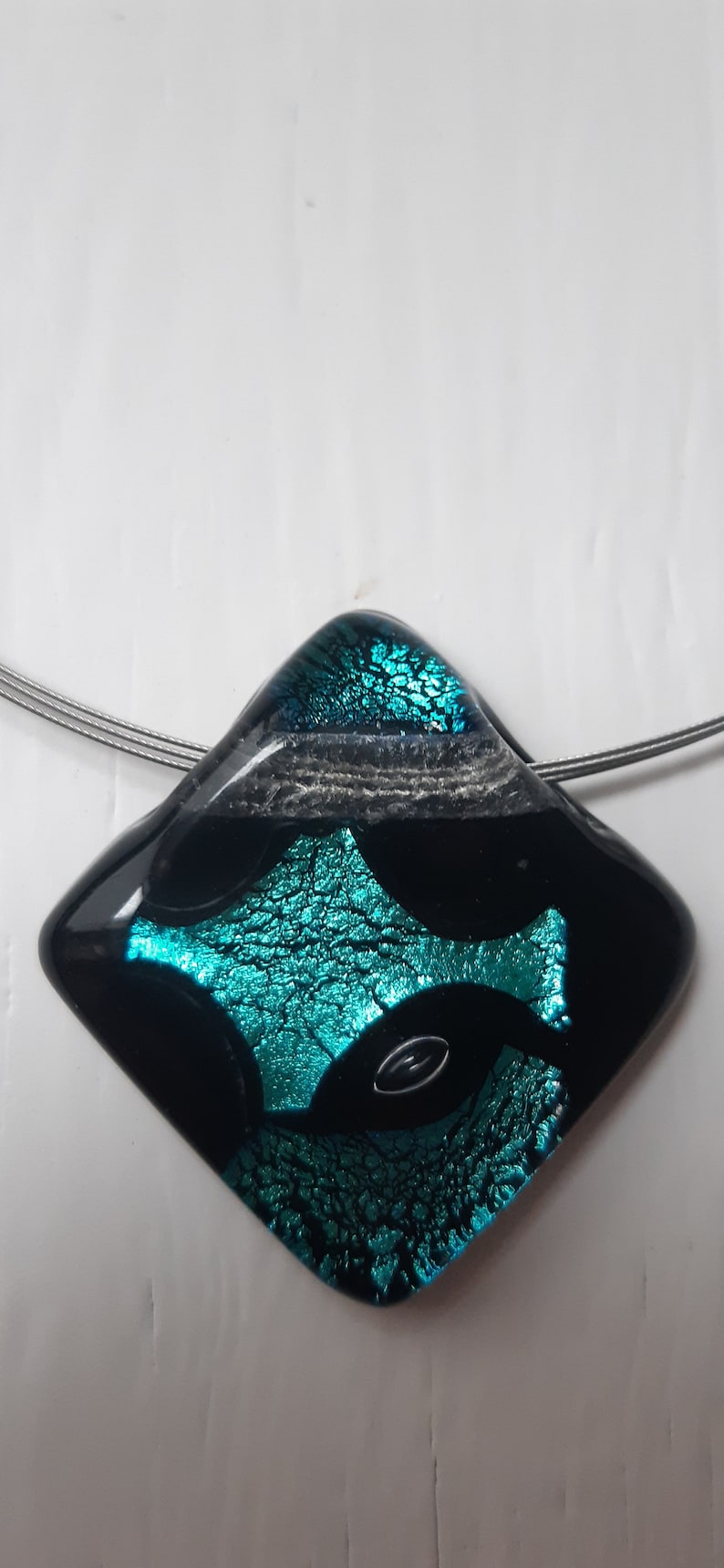 May include: A black and teal glass pendant with a diamond shape. The pendant has a silver wire attached to it.
