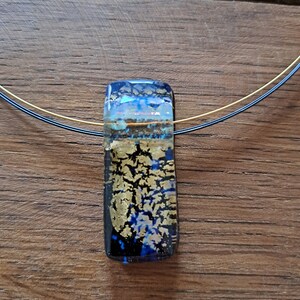 May include: A rectangular pendant necklace with a dichroic glass pendant. The pendant features a mix of blue, gold, and clear glass, with a gold leaf design. The necklace has a thin, multi-strand wire. The background is a wooden surface.