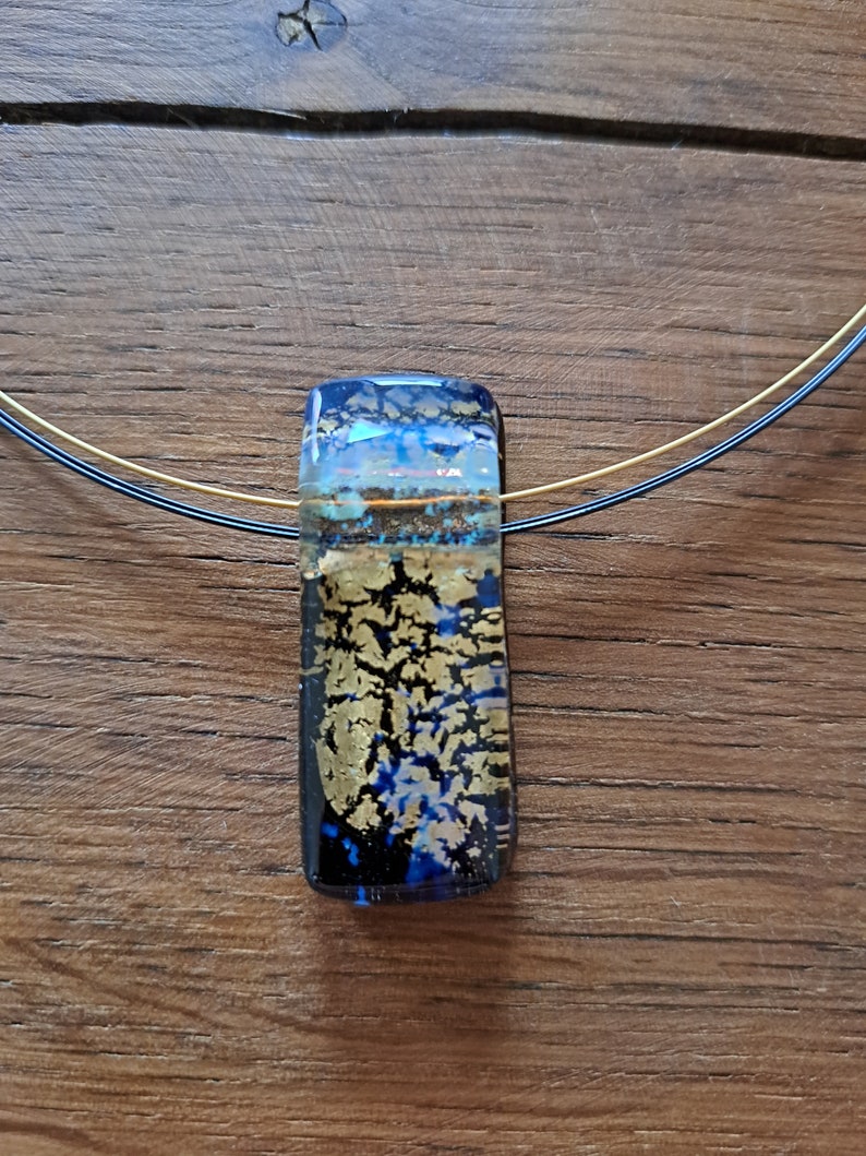 May include: A rectangular pendant necklace with a dichroic glass pendant. The pendant features a mix of blue, gold, and clear glass, with a gold leaf design. The necklace has a thin, multi-strand wire. The background is a wooden surface.