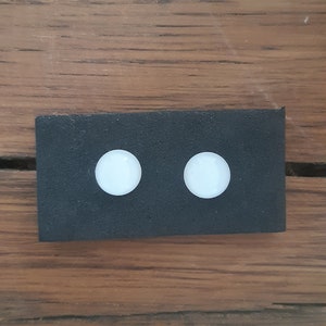 May include: A black rectangular object with two white, circular objects evenly spaced on its surface. The background is a wooden surface with a brown colour and visible wood grain.