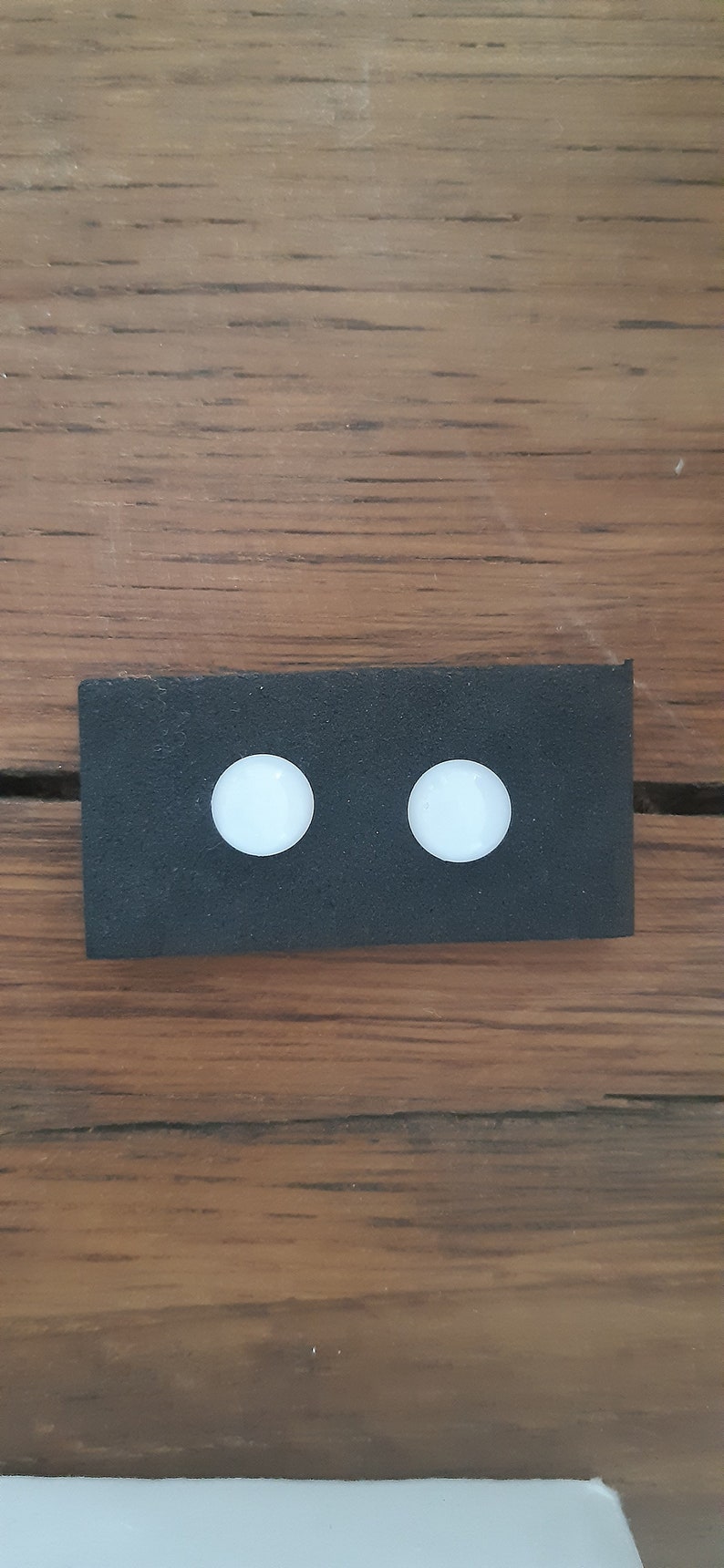 May include: A black rectangular object with two white, circular objects evenly spaced on its surface. The background is a wooden surface with a brown colour and visible wood grain.
