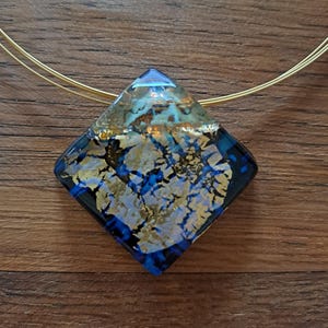 May include: A gold and blue glass pendant necklace with a square-shaped pendant. The pendant has a marbled design with gold and blue swirls.
