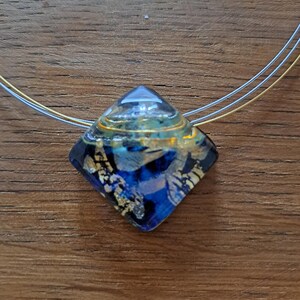 May include: A square-shaped glass pendant necklace with a blue, gold and black abstract design. The pendant is suspended from a thin silver chain.