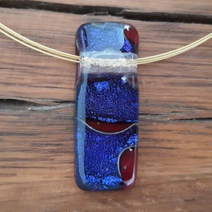 May include: A blue and red glass pendant with gold accents hangs from a gold chain. The pendant has a textured surface and a layered design.