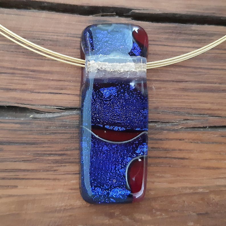 May include: A blue and red glass pendant with gold accents hangs from a gold chain. The pendant has a textured surface and a layered design.
