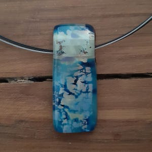 May include: A rectangular pendant necklace with a blue and white abstract design. The pendant is made of glass and has a glossy finish. The necklace cord is a thin, dark gray wire.
