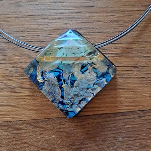 May include: A square-shaped glass pendant necklace with a blue, gold, and white abstract design. The pendant is attached to a thin black cord.