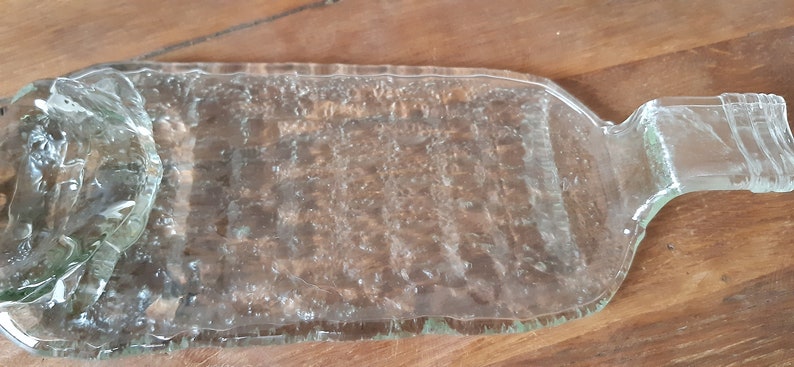 May include: A clear glass bottle that has been repurposed into a serving tray. The bottle has a textured surface and a handle on one end.