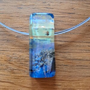 May include: A rectangular pendant with a blue, white, and yellow design, suspended on a thin, clear cord. The pendant has a layered appearance, with a textured, abstract pattern. The overall aesthetic is reminiscent of an underwater scene.