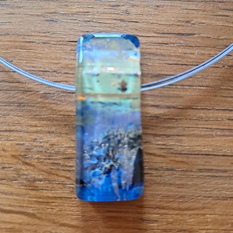 May include: A rectangular pendant with a blue, white, and yellow design, suspended on a thin, clear cord. The pendant has a layered appearance, with a textured, abstract pattern. The overall aesthetic is reminiscent of an underwater scene.