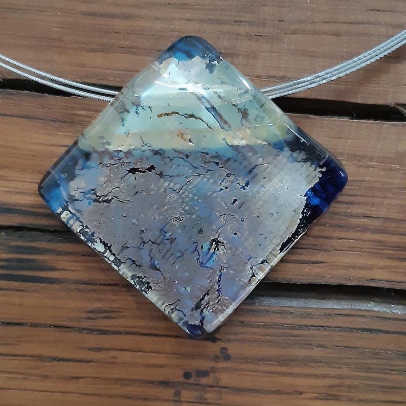May include: A square glass pendant with a silver and blue design. The pendant is on a silver chain.