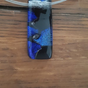 K&ouml;nnte beinhalten: Ein blauer und schwarzer Glasanh&auml;nger mit einer silbernen Kette. Der Anh&auml;nger hat ein einzigartiges, abstraktes Design mit Wirbeln aus blauem und schwarzem Glas.